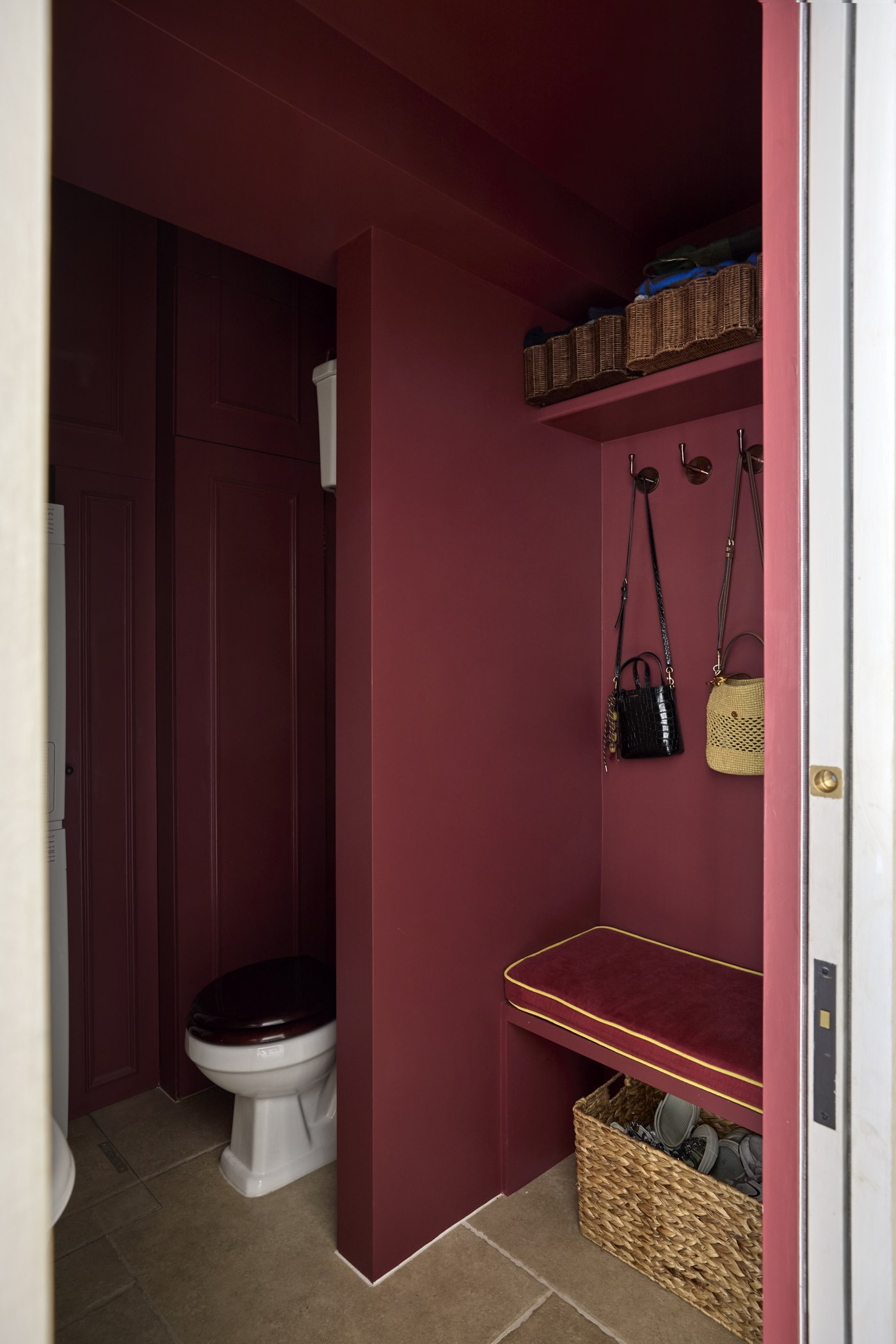 Honeywell Road SW11 — cloakroom in the same deep red, with an upholstered bench, coat hooks and a high shelf over stone floor tiles