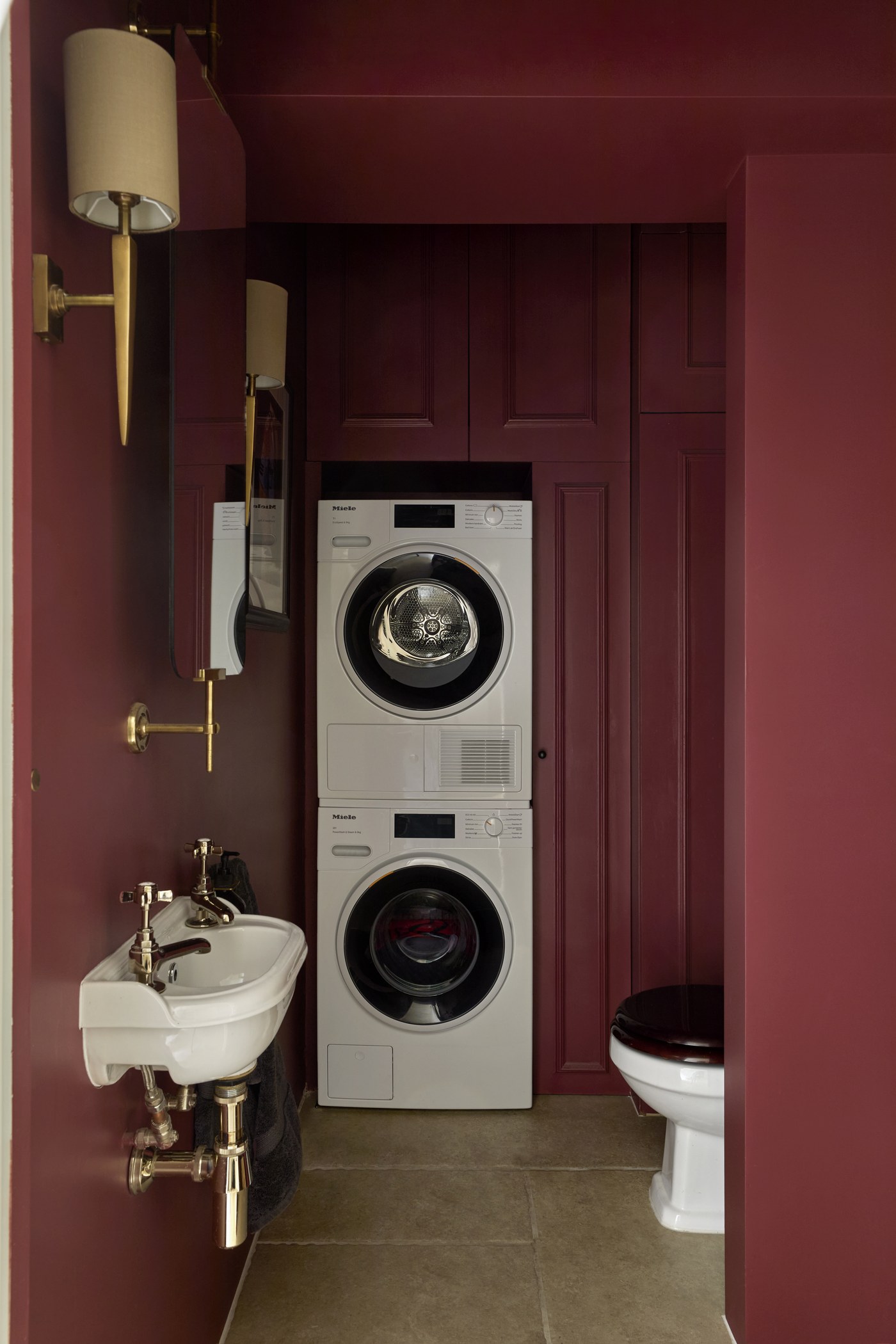 Honeywell Road SW11 — utility room finished in deep red, with a stacked washer and dryer behind panelled joinery, a small basin and brass wall lights